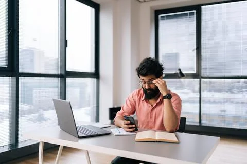 Remote view of puzzled Indian freelancer male in glasses using smartphone Stock Photos