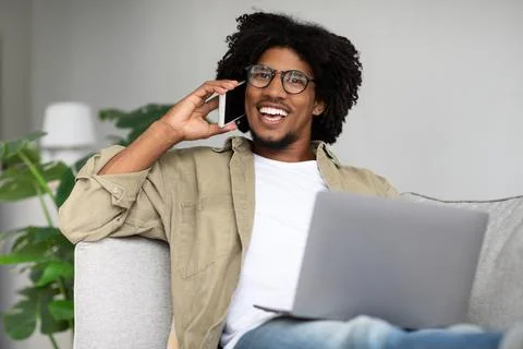 Remote Work. Black Guy Talking On Cellphone And Using Laptop At Home Stock Photos