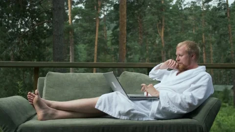 Remote work, a man in a robe works at a laptop on the couch in the woods 스톡 동영상 140661024