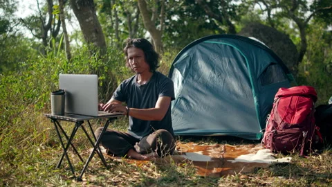 Remote worker using a laptop beside a camping tent in a grassy forest meadow Stock Footage 341767424