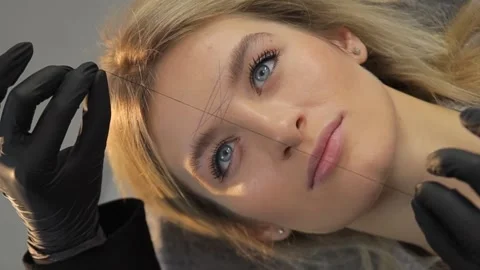 Removing unwanted hair from the eyebrows with a thread, a cosmetologist does Stock Footage 264073120
