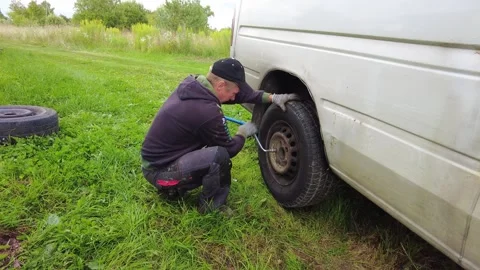 Replacing a flat tire on a van while situated in a grassy area under the warm ra Stock Footage 345962544