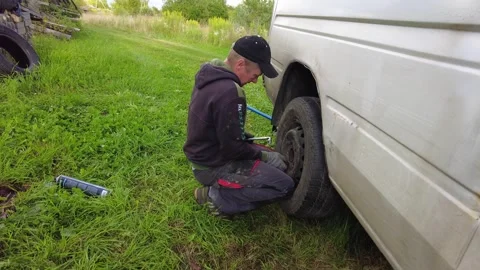 Replacing a flat tire on a van while situated in a grassy field during the dayli Stock Footage 347024302