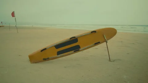 Rescue Board Standing Upright on Sandy Beach with Warning Flag by the Ocean Stock Footage 344040034