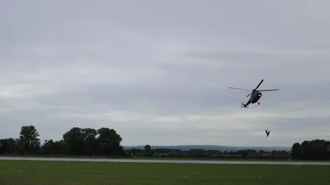 Rescue helicopter hovers in cloudy sky above airfield as crew in red suit dangle Stock Footage 311662099
