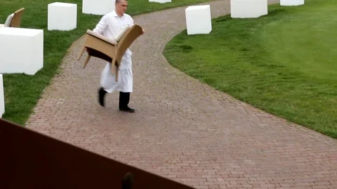 A restaurant worker in white uniform carries a chair across the lawn Stock Footage 89296623