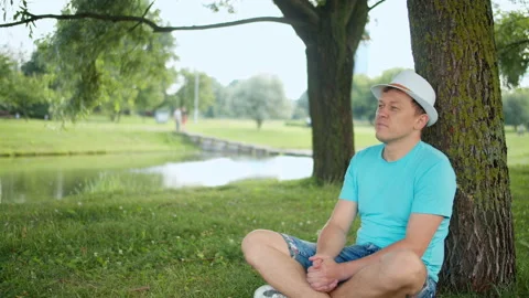 Resting young man sitting under a tree in a city park, camera tracking Stock Footage 139289013