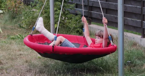Restless kid with leg in cast lying on garden nest swing Video stock 298863423