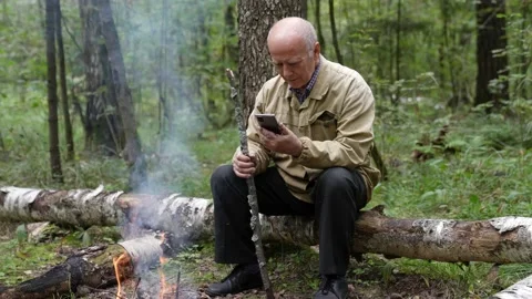 A retired man in the forest by the fire sits on a log and uses a phone to Stock-Footage 159640089