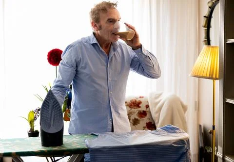 Retired man having a coffee while taking a break from ironing Stock Photos