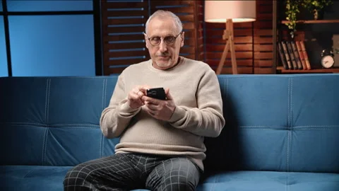 Retired man sitting on sofa in room and using mobile app on smartphone, portrait Stock Footage 264086483