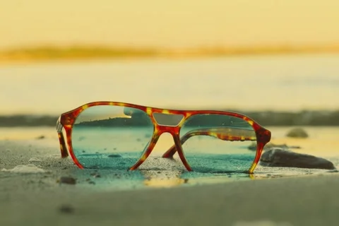 Retro tortoise sunglasses on beach with wave crashing through lenses Stock Footage 332399569