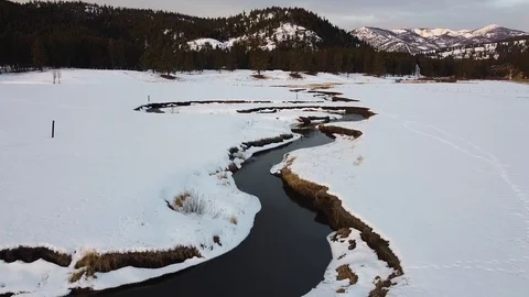 Reverse overhead dolly shot over snow covered field with winding creek, fores Stock Footage 125384589
