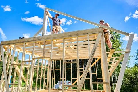 Ridge purlin mounted, young workers position rafters above it at constructi.. Stock Photos