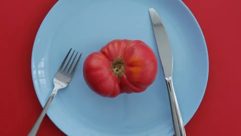 Ripe red tomato with knife and fork on a blue plate with a red background. Stock Footage 344567836