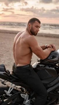 Ripped man with muscular torso on his black sports motorcycle on desolate beach Stock Photos