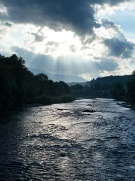 River and dramatic sky with sun rays through dark clouds in mountains Stock Photos