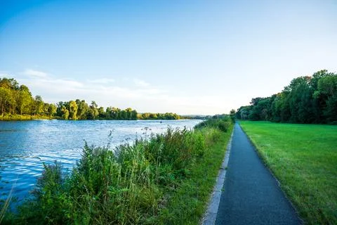 River embankment with a paved walking path between blue water and a green meadow Stock Photos