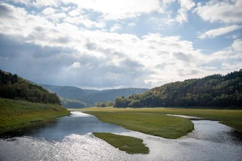 River Flows Through a Grassy Valley with Green Hills and Cloudy Sky Stock Photos