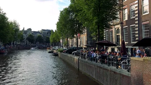 A river in a traditionally looking town, people sitting under parasols, trees Stock Footage 73958658