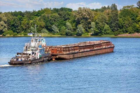 A river tugboat with an empty rusty barge goes crosses a wide river. Stock Photos