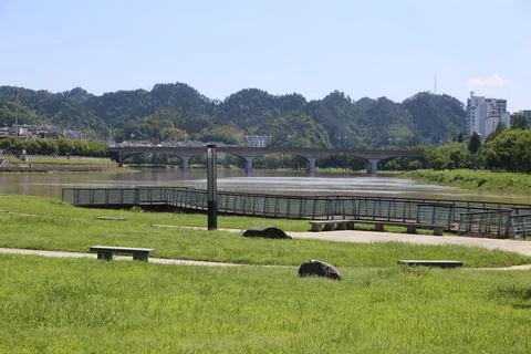 Riverside lawn and viewing platform overlook a bridge, wooded hills, and br.. Stock Photos