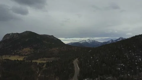 A road among thick trees winds through mountains leading to a large snowy peak Stock Footage 167675272