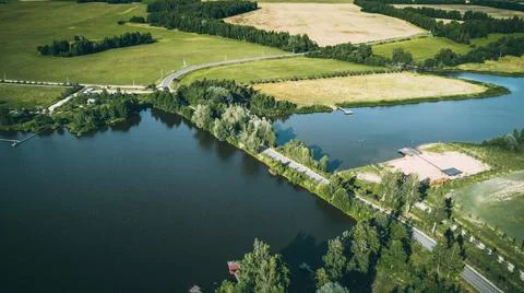The road among the trees runs between two lakes, around green fields Stock Photos
