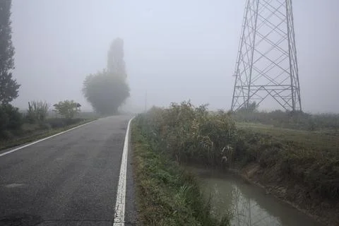 Road bordered by trenches with a pylon in a field by its edge on a foggy da.. Stock Photos
