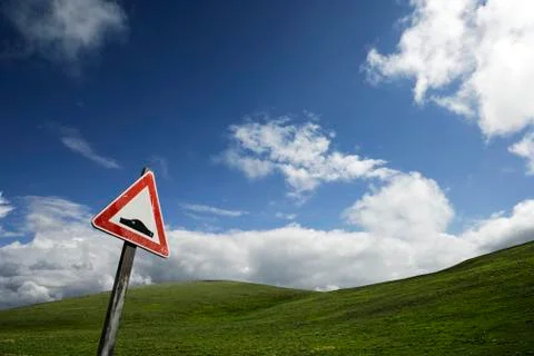 Road sign warning about a bumpy road, in front of a green hill under a blue s Stock Photos