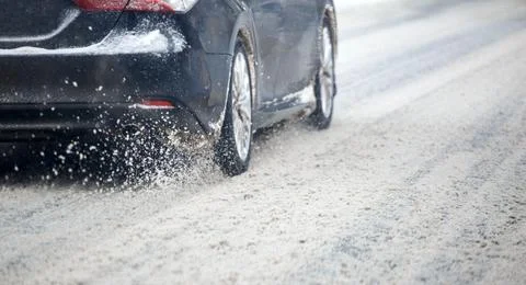 Road snow flinders flow from wheels of black car moving fast in daylight city 库存照片