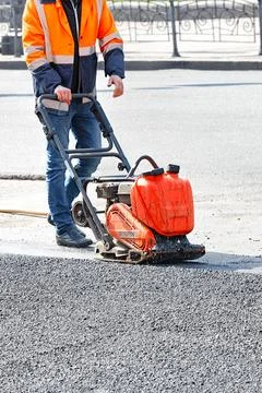 A road worker in an orange jacket compacts a patch of asphalt with a petrol v Stock Photos