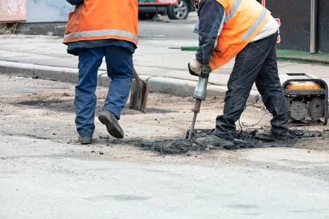 Road workers in orange vests break up old asphalt with an electric jackhammer Stock Photos