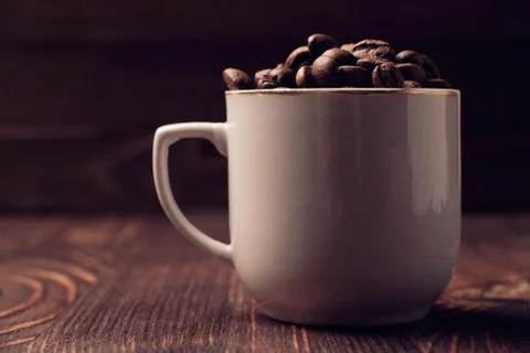 Roasted coffee beans in a white cup on a burnt wooden background. Toned Stock Photos