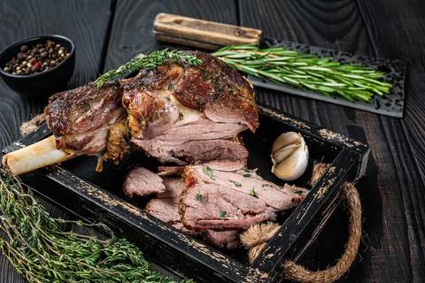 Roasted mutton lamb leg sliced in a wooden tray with meat cleaver. Black wooden Stock Photos