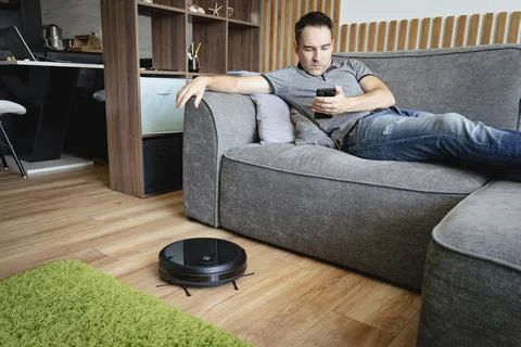 Robot vacuum cleaner cleaning the living room. man enjoy rest, sitting on sof 写真素材