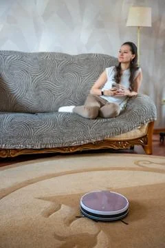 Robot vacuum cleaning the floor while a young woman relaxes on the couch in the Stock Photos