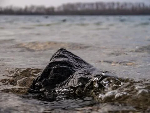 Rock inside a lake, getting hit by a wave, with a tree line in the background Stock Photos