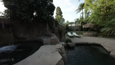 Rock waterfall flowing into a backyard pool beside lounge chairs Stock Footage 345543308
