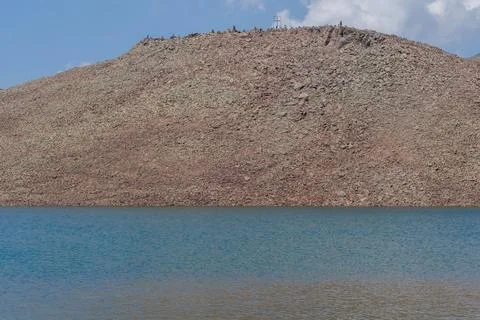 Rocky hillside with sparse vegetation reflected in calm blue lake under a clear Stock Photos