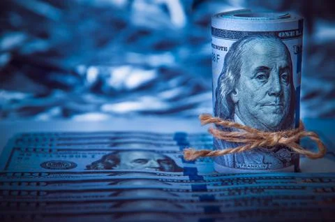 A roll of dollars on the background of scattered hundred dollar bills in blue Stock Photos