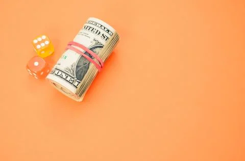 A rolled-up stack of dollars next to colorful dice on a pastel background wit Stock Photos