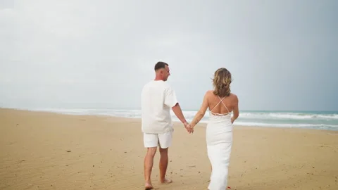 Romantic couple dressed in white walking hand in hand along sandy beach bes.. Vídeo Stock 345511732