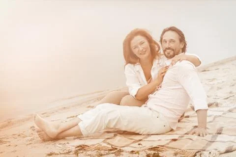 Romantic couple looks at camera while sitting on sandy coast. Happy woman and Stock Photos
