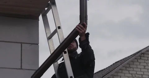 A roofer installs a gutter system. The focused craftsman attaches a downspout to Video stock 322934310