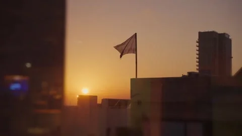 Rooftop flag gently waving at sunset above a modern city skyline, warm golden 動画素材 332707821