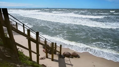 Rough sea waves crashing onto sandy beach near wooden staircase on sunny day Stock Footage 339577702