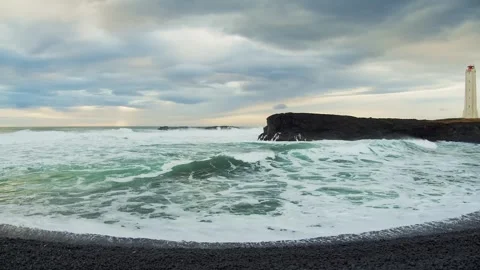Rough waves crashing on a black sand beach with a lighthouse in Iceland. 스톡 동영상 320202035