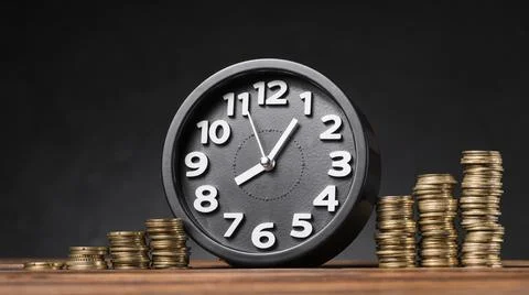 Round clock between the increasing coins on wooden desk against black backdrop Stock Photos