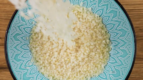 Round grain rice is poured into a ceramic blue bowl against a wooden oak board.  Video stock 304395324